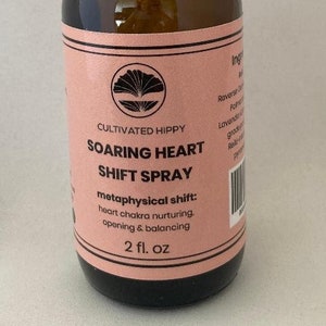 May include: A brown glass bottle with a black spray top and a pink label. The label reads "Cultivated Hippy Soaring Heart Shift Spray" and "metaphysical shift: heart chakra nurturing opening & balancing 2 fl. oz".