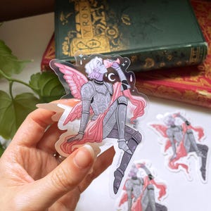 Op de afbeelding: Transparante sticker met een zittende fee-achtige figuur in harnas met roze vleugels en een roze sjerp. De figuur houdt een drankje vast. Twee extra stickers zijn zichtbaar. De achtergrond bevat een groen boek met gouden letters.