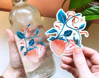 Può includere: Una bottiglia di vetro trasparente con tappo nero e un adesivo a forma di fragola. L'adesivo presenta un'illustrazione ad acquerello di fragole, foglie blu e nastri rossi. Un secondo adesivo a forma di fragola è tenuto in mano.
