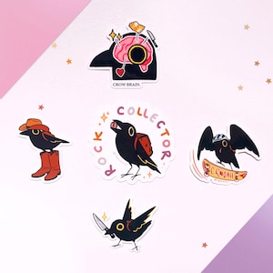 May include: A selection of quirky crow stickers. One sticker shows a crow with a brain graphic and the text "CROW BRAIN." Other stickers depict crows with cowboy boots, a backpack, a skateboard, and a knife. The stickers are on a white background.
