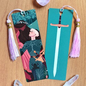 May include: Two rectangular paper bookmarks with light pink tassels. One bookmark features an illustration of a person in dark green armor facing a person with long dark hair. The other bookmark displays a long sword on a solid teal background.