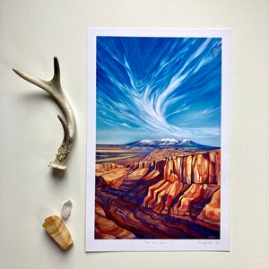May include: A landscape art print featuring a canyon scene with a vibrant blue sky filled with swirling clouds. The canyon walls are shades of red and brown. A deer antler and two stones are next to the print.