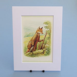 Könnte beinhalten: Gerahmter Druck eines Fuchses in einem braunen Mantel, der sich mit einem Wanderstock an einer Steinmauer abstützt. Die Illustration hat einen Vintage-Look, vor dem Hintergrund grüner Felder und Hügel. Der Druck ist in einem weißen Passepartout montiert.