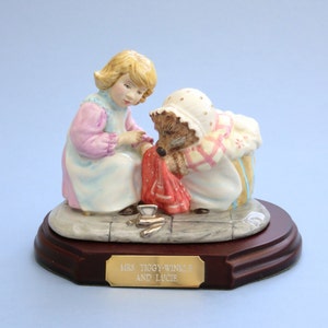 Może przedstawiać: Ceramiczna figurka przedstawiająca Panią Tiggy-Winkle i Lucie. Figurka przedstawia dziewczynkę w różowo-niebieskiej sukience i jeża w białym kapturze i fartuchu. Podstawa to ciemne drewno ze złotą plakietką z napisem "MRS TIGGY-WINKLE AND LUCIE."
