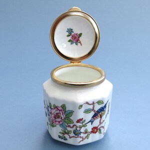 May include: A white porcelain jar with a gold rim and lid, adorned with floral and bird patterns. The lid is open, displaying a small floral design. The jar has a hexagonal shape and is set against a light blue backdrop.