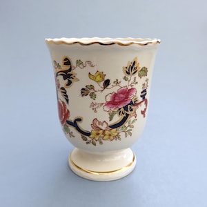 May include: Cream ceramic vase with a scalloped rim and pedestal base, decorated with pink, yellow, and red floral patterns. The vase has gold trim and dark blue accents.