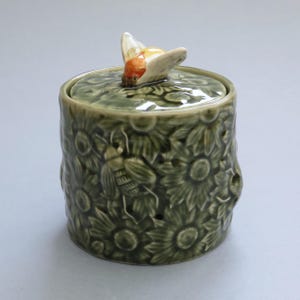Pode incluir: Um pote cilíndrico de cerâmica com esmalte verde-oliva brilhante, apresentando um padrão em relevo de girassóis e abelhas. A tampa é rematada com uma figura de abelha esculpida e pintada à mão com detalhes em laranja e branco.