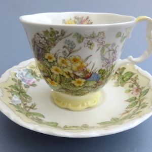 May include: A fine bone china teacup and saucer set. The white cup and saucer are decorated with a detailed floral pattern, including yellow primroses and a small cottage scene. The cup has a decorative handle.