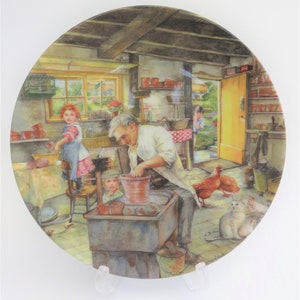 May include: Decorative ceramic plate with a detailed watercolour scene of a potting shed. The image shows a man working with clay, children, chickens, and cats. The plate has a glossy finish and is displayed on a clear stand.