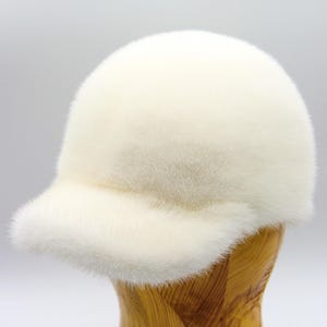 May include: A white fur baseball cap with a curved brim. The cap is displayed on a wooden hat stand against a white background. The fur appears soft and plush.