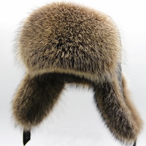 May include: A brown fur hat with ear flaps. The hat features a rounded crown and ear flaps that extend downwards. The fur is a mix of brown and tan colours. The ear flaps are lined with a darker material and have black straps.