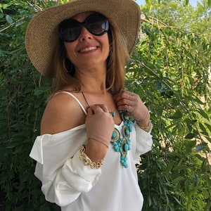 Puede incluir: Una mujer con una blusa blanca con un collar de declaraci&oacute;n turquesa y dorado, un sombrero de paja de ala ancha y gafas de sol. Est&aacute; de pie frente a un fondo verde frondoso.