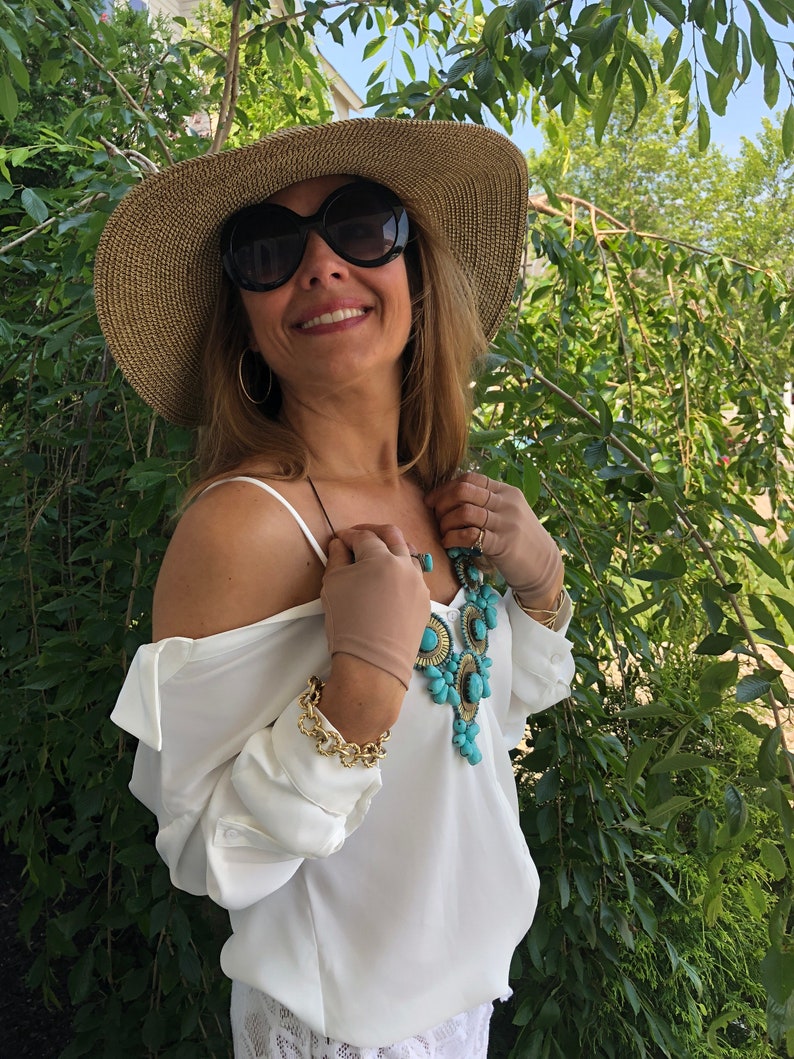Puede incluir: Una mujer con una blusa blanca con un collar de declaraci&oacute;n turquesa y dorado, un sombrero de paja de ala ancha y gafas de sol. Est&aacute; de pie frente a un fondo verde frondoso.