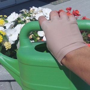 Puede incluir: Una mano con un guante beige sin dedos sostiene el asa de una regadera verde. La regadera est&aacute; junto a una maceta llena de flores de colores.