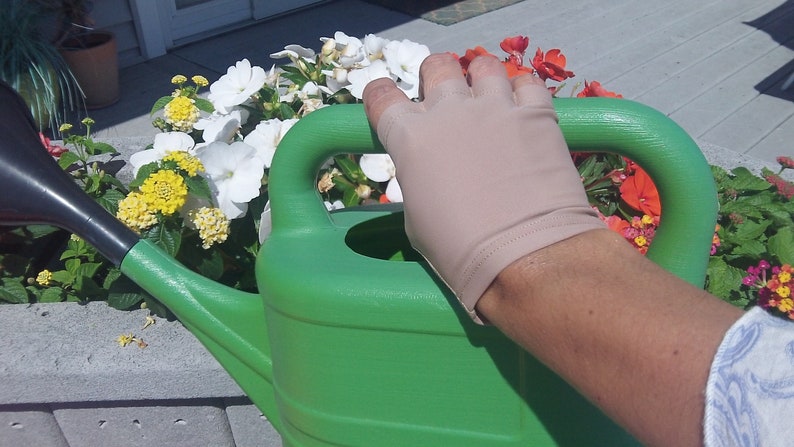 Puede incluir: Una mano con un guante beige sin dedos sostiene el asa de una regadera verde. La regadera est&aacute; junto a una maceta llena de flores de colores.