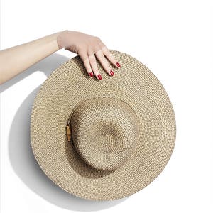 Puede incluir: Un sombrero de paja de ala ancha con un patr&oacute;n tejido marr&oacute;n y negro. El sombrero est&aacute; sostenido por una mano con esmalte de u&ntilde;as rojo.