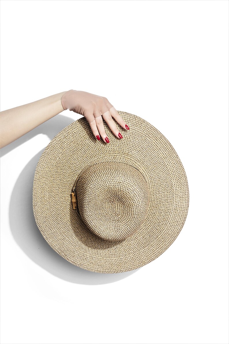 Puede incluir: Un sombrero de paja de ala ancha con un patr&oacute;n tejido marr&oacute;n y negro. El sombrero est&aacute; sostenido por una mano con esmalte de u&ntilde;as rojo.