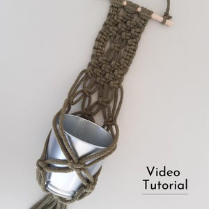 Puede incluir: Un macetero de macramé verde con una maceta de metal plateada colgando de él. El macetero está sujeto a una varilla de madera. El texto "Video Tutorial" está en la parte inferior de la imagen.