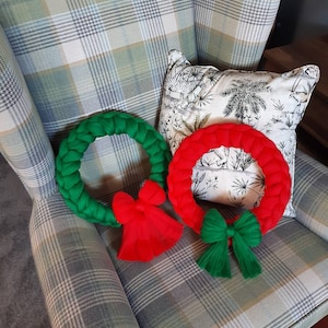 Könnte beinhalten: Zwei Weihnachtskränze aus Garn, einer grün und einer rot, mit passenden Schleifen. Die Kränze liegen auf einem karierten Sessel.