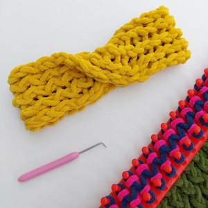 May include: A mustard yellow, chunky knit headband with a twisted design. A pink crochet hook and a red, pink, and blue knitting loom are also visible. The headband is made of a soft, textured yarn.