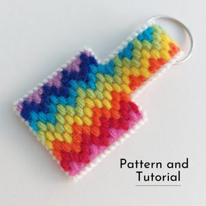 May include: A handmade, rainbow-colored arrow-shaped keychain. The keychain is made of stitched yarn in a chevron pattern, with a white border and a silver key ring. The text "Pattern and Tutorial" is printed on the bottom right.
