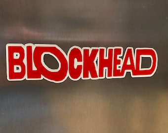 May include: A red and white sticker with the word "BLOCKHEAD" in a bold, rounded font. The text is red with a white outline, and it is affixed to a metallic surface.