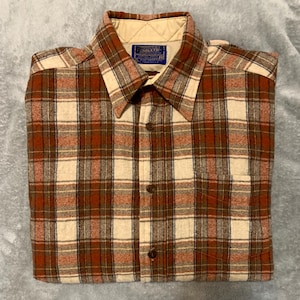 May include: A folded, button-down shirt with a plaid pattern in shades of brown, cream, and rust. The shirt has a collar and a pocket on the front. A small label is visible near the collar.