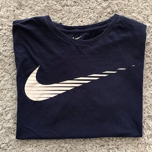May include: Navy blue t-shirt with a white Nike logo. The logo is a stylized check mark with horizontal lines. The shirt is folded neatly on a gray, textured surface.