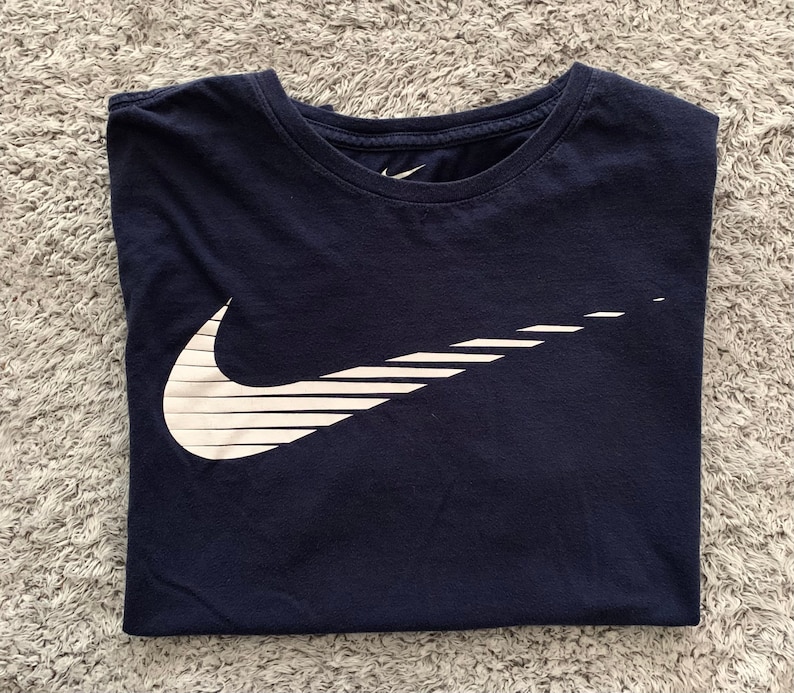 May include: Navy blue t-shirt with a white Nike logo. The logo is a stylized check mark with horizontal lines. The shirt is folded neatly on a gray, textured surface.
