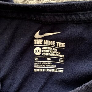 May include: Close-up of a navy blue athletic t-shirt. The Nike logo and "THE NIKE TEE" are visible in white. The shirt has a tag with text including "ATHLETIC CUT" and "MADE IN HONDURAS". The website "NIKEBETTERWORLD.COM" is also printed on the tag.