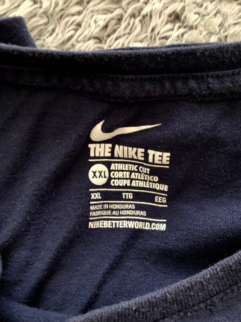May include: Close-up of a navy blue athletic t-shirt. The Nike logo and "THE NIKE TEE" are visible in white. The shirt has a tag with text including "ATHLETIC CUT" and "MADE IN HONDURAS". The website "NIKEBETTERWORLD.COM" is also printed on the tag.