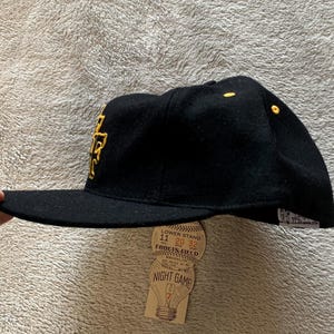May include: Black baseball cap with a gold embroidered logo. The cap has a flat brim and a tag that reads "Ebbets Field Night Game". The cap is a classic style, perfect for sports fans.