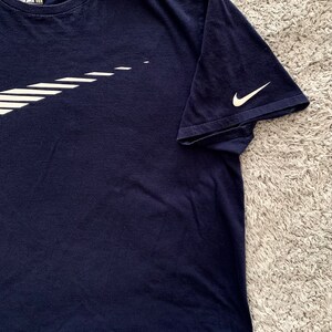 May include: Navy blue t-shirt with a white Nike logo on the front and sleeve. The logo is a stylized swoosh design. The shirt is made of a soft material and has a crew neck.