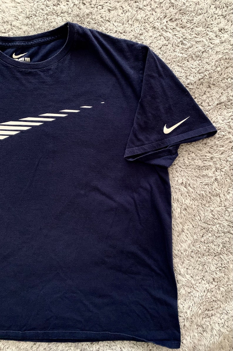 May include: Navy blue t-shirt with a white Nike logo on the front and sleeve. The logo is a stylized swoosh design. The shirt is made of a soft material and has a crew neck.