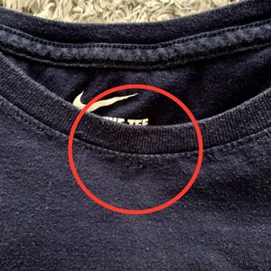 May include: Close-up of a navy blue crew neck t-shirt. The white Nike logo is visible on the upper left chest. The text "NIKE TEE" is printed in white below the logo. The shirt is made of a soft, textured fabric.