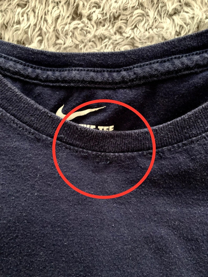May include: Close-up of a navy blue crew neck t-shirt. The white Nike logo is visible on the upper left chest. The text "NIKE TEE" is printed in white below the logo. The shirt is made of a soft, textured fabric.
