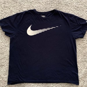 May include: Navy blue t-shirt featuring a large white Nike swoosh logo on the front. The logo has a stylized, dynamic design with horizontal lines. The Nike logo is also on the left sleeve.