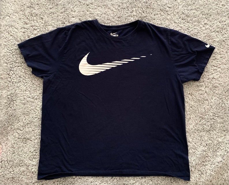 May include: Navy blue t-shirt featuring a large white Nike swoosh logo on the front. The logo has a stylized, dynamic design with horizontal lines. The Nike logo is also on the left sleeve.