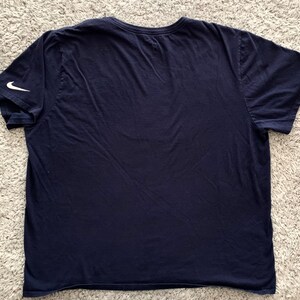 May include: Navy blue t-shirt with a crew neck and short sleeves. A white Nike swoosh logo is visible on the left sleeve. The shirt is made of a soft, likely cotton, material.