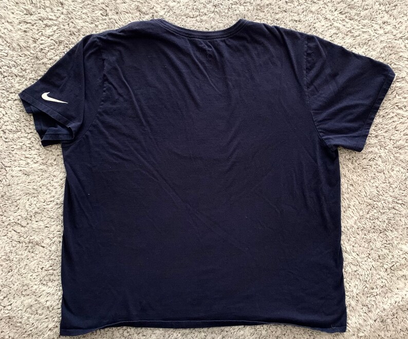 May include: Navy blue t-shirt with a crew neck and short sleeves. A white Nike swoosh logo is visible on the left sleeve. The shirt is made of a soft, likely cotton, material.