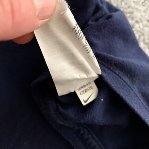 May include: Close-up of a navy blue garment with a white tag attached. The tag has text and a Nike logo. The tag reads "472303-2XL".