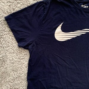 May include: Navy blue t-shirt with a white Nike logo on the front. The logo is a stylized check mark with horizontal lines. The shirt has short sleeves and a crew neck. The Nike logo is on the upper left chest.