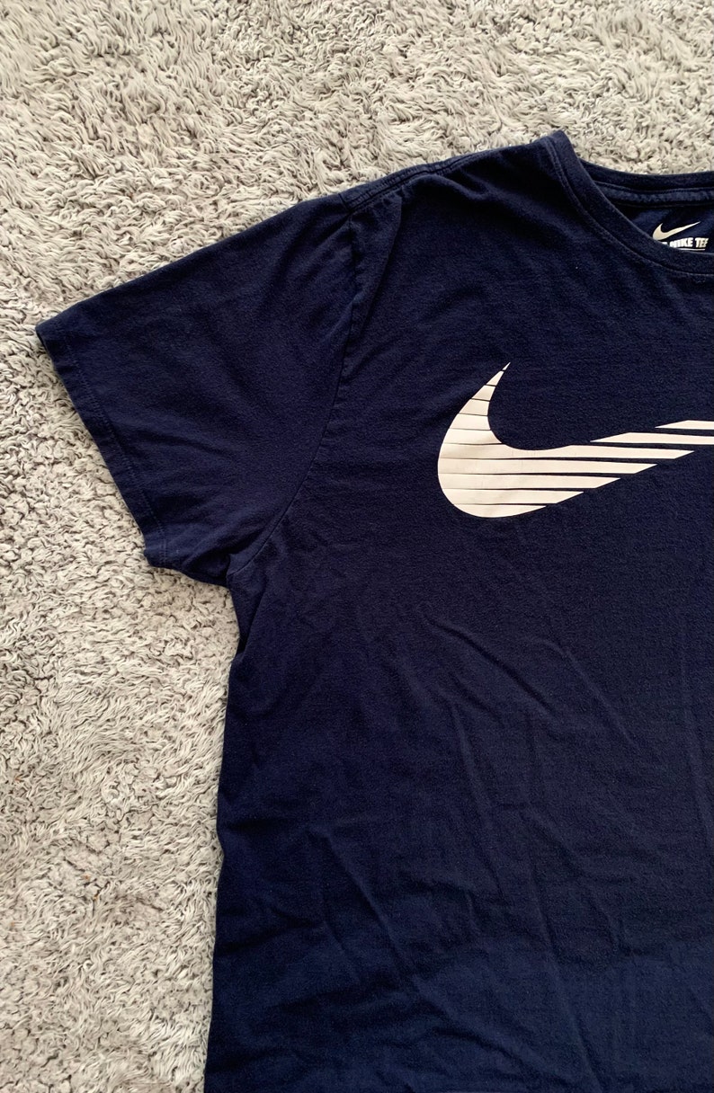May include: Navy blue t-shirt with a white Nike logo on the front. The logo is a stylized check mark with horizontal lines. The shirt has short sleeves and a crew neck. The Nike logo is on the upper left chest.