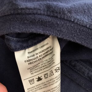 May include: Close-up of a navy blue garment tag with text in English, French, and Spanish. The tag includes washing instructions and the text "MADE IN HONDURAS". The garment appears to be made of cotton.