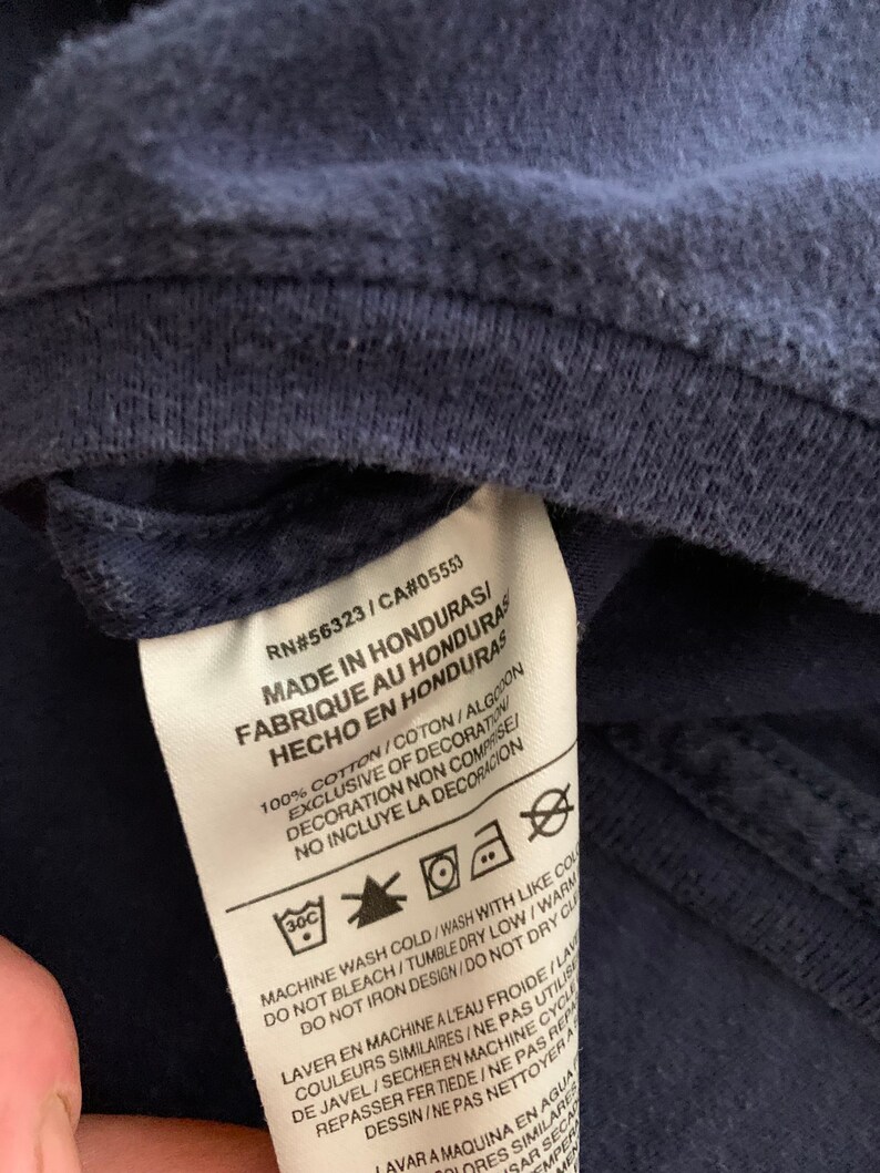 May include: Close-up of a navy blue garment tag with text in English, French, and Spanish. The tag includes washing instructions and the text "MADE IN HONDURAS". The garment appears to be made of cotton.