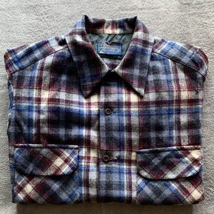 May include: A blue, red, and white plaid shirt with two front pockets and a collared neckline. The shirt has two buttons and a label on the collar.