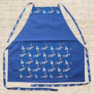 Puede incluir: Un delantal azul con un bolsillo grande en la parte delantera. El bolsillo está forrado con una tela blanca con un patrón repetido de pelotas de béisbol y las palabras "LA Dodgers". El delantal tiene correas ajustables.