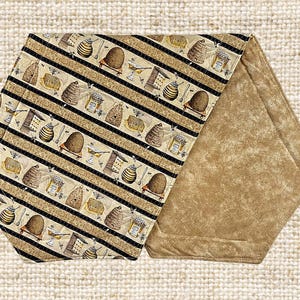 Könnte beinhalten: Ein dekoratives, sechseckiges Küchenutensil mit einem Bienenmotiv. Die Oberseite zeigt ein sich wiederholendes Muster aus Bienenstöcken, Bienen und Honigtöpfen auf beigem Hintergrund mit schwarzen Streifen. Die Unterseite ist einfarbig beige.