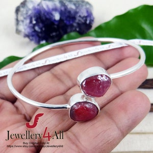 May include: A silver bangle bracelet with two red ruby gemstones. The bracelet is open and has a simple design. The rubies are rough cut and have a natural, organic look.