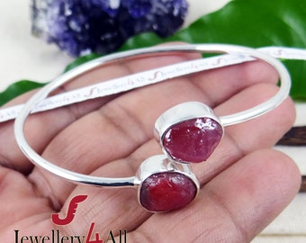 May include: A silver bangle bracelet with two red ruby gemstones. The bracelet is open and has a simple design. The rubies are rough cut and have a natural, organic look.